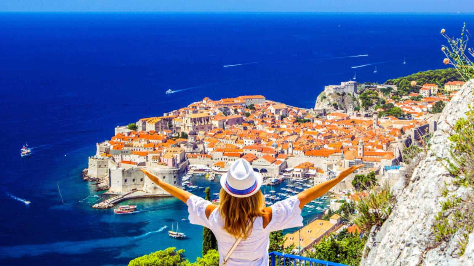 Happy woman enjoys view of old town (medieval Ragusa) and Dalmatian Coast of Adriatic Sea in Dubrovnik. Blue sea with white yachts, beautiful landscape, aerial view, Dubrovnik, Croatia