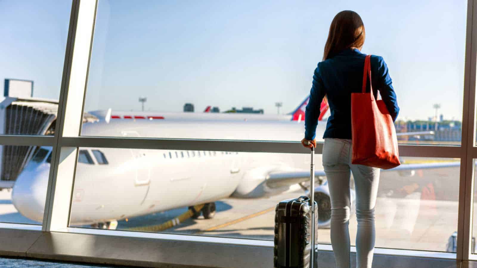 Travel tourist standing with luggage watching sunset at airport window. Unrecognizable woman looking at lounge looking at airplanes while waiting at boarding gate before departure. Travel lifestyle.