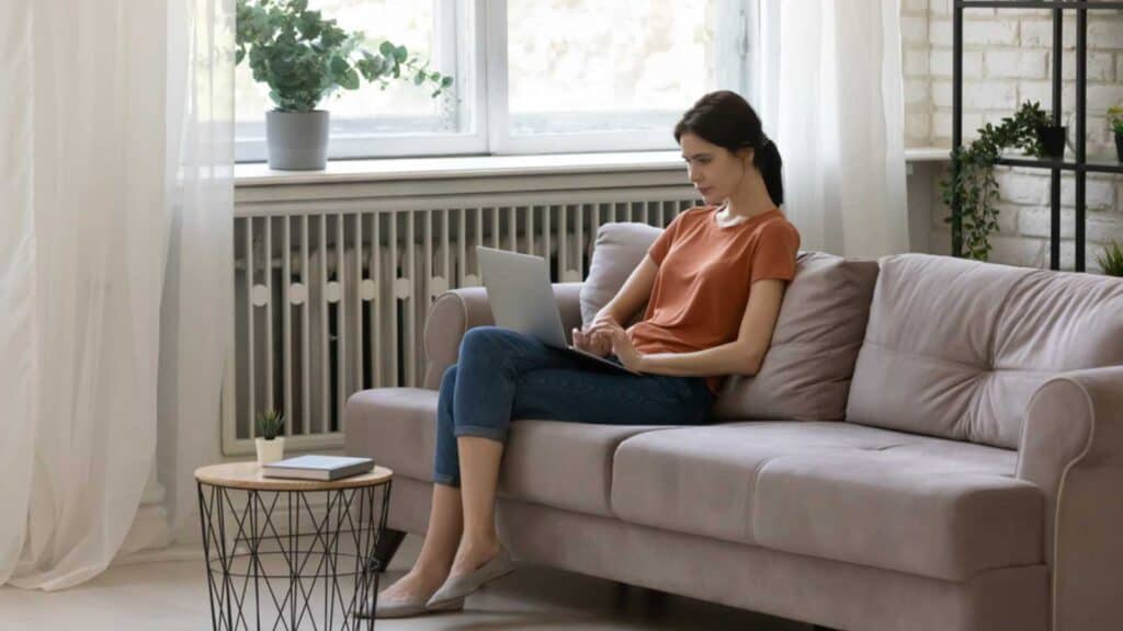 Online activity. Calm young woman sitting with crossed legs on cozy sofa in living room holding laptop on knees. Tranquil female focused on pc screen shopping chatting online surfing web writing email