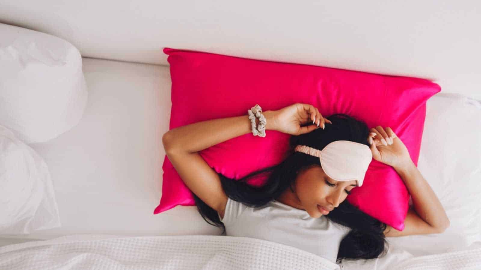 Beautiful Indonesian girl lying on a bright pink pillow with a silk pillowcase and a sleep mask. View from above