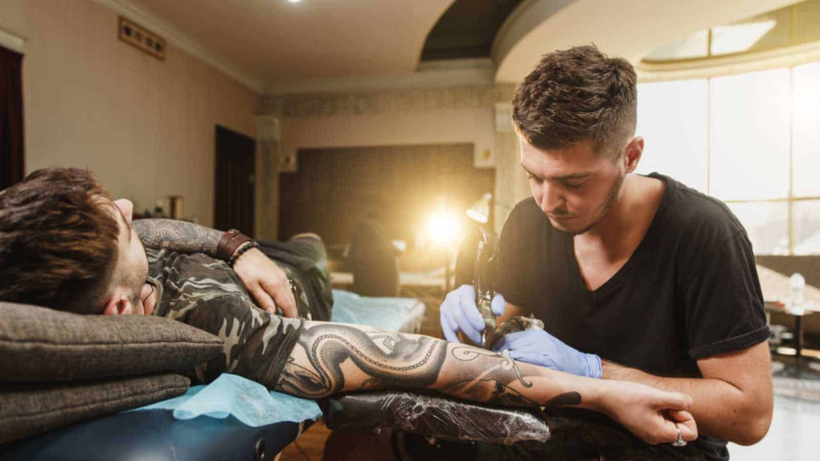 A professional tattooer artist doing picture on hand of man by machine black ink from a jar. Tattoo art on body. Equipment for making tattoo art. Master makes tattooed in light studio.
