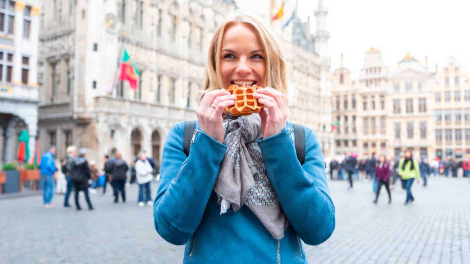 A beautiful young woman is standing on the square Grand Place in Brussels and is going to eat a delicious waffle - the national sweetness of Belgium.