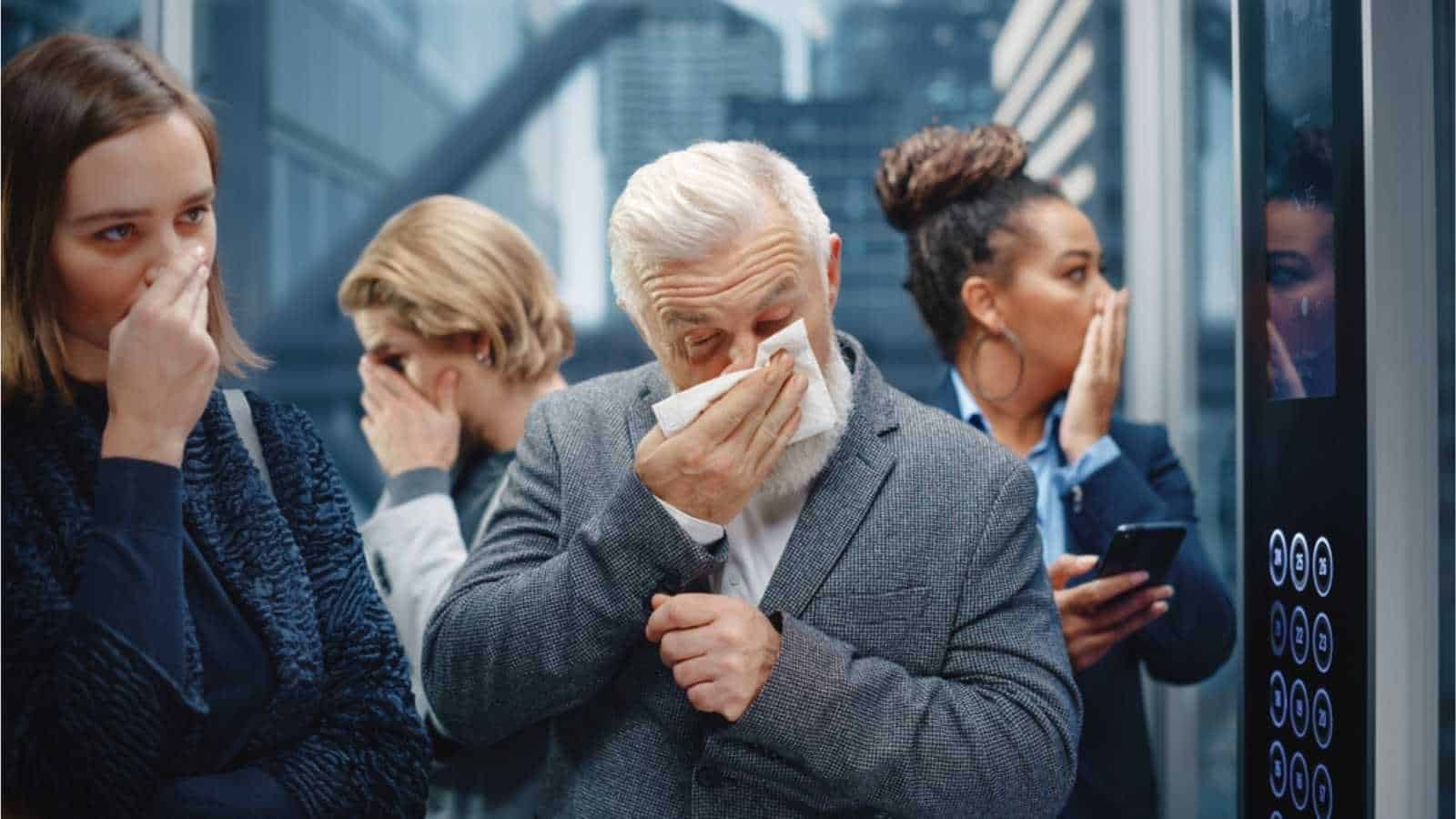 Middle Aged Man Sneezes in a Crowded Glass Elevator in a Modern Office Building. Businessman Covers His Face, but Other People Are Afraid to Catch the Virus and Microbes in a Lift.
